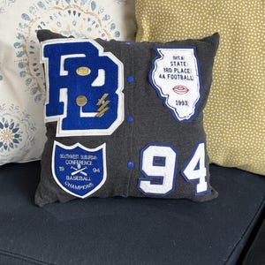 May include: A gray pillow with blue and white patches. The patches include a large blue "B", a patch with the text "IHSA STATE 3RD PLACE 4A FOOTBALL 1993", a patch with the text "SOUTHWEST SUBURBAN CONFERENCE 19 BASEBALL CHAMPIONS 94", and a large white "94".