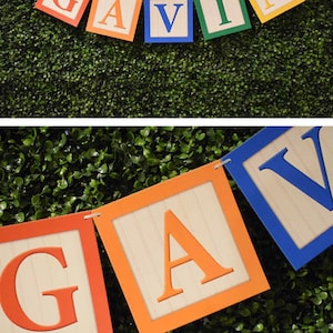 K&ouml;nnte beinhalten: Ein Banner mit dem Namen "GAVIN", der in bunten Blockbuchstaben geschrieben ist. Jeder Buchstabe befindet sich auf einem separaten Block mit Holzmaserung. Die Bl&ouml;cke sind mit einer Schnur aneinandergereiht.