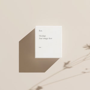 Pu&ograve; includere: Mockup di scatola quadrata bianca con il testo "Box Mockup Your image here PSD" su uno sfondo beige con l'ombra di una pianta.