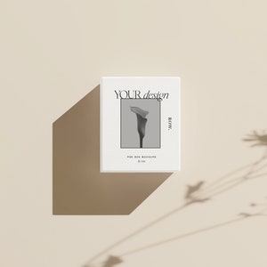 Pu&ograve; includere: Mockup di scatola bianca con una foto in bianco e nero di un fiore e il testo "YOUR design ROW." La scatola &egrave; su uno sfondo beige.
