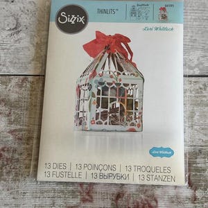 May include: A Sizzix Thinlits die set by Lori Whitlock in a clear plastic package. The packaging features a graphic of a light blue, floral-patterned, lantern-shaped box topped with a red ribbon. The text reads Sizzix, Thinlits, 661095, and 13 dies.