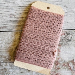 May include: A roll of dusty rose lace trim with a delicate circular pattern. The lace is folded and displayed on a brown card.