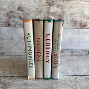 May include: A collection of four vintage books with white spines and coloured edges. Titles visible are "Automobiles", "Lichens", "Geology", and "The Observer's Book of Ships". The books are set against a light-coloured, weathered wooden background.