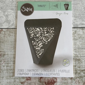 May include: A Sizzix Thinlits die set, model 664447 by Georgia Evans, packaged in a clear plastic sleeve. The packaging features a dark grey triangular box with a decorative floral cutout pattern, the Sizzix logo, and text indicating 2 dies.
