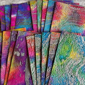 May include: A selection of colourful handmade paper sheets. The sheets display vibrant hues of pink, purple, blue, yellow, and green with silver accents. The paper has a textured, marbled appearance, suggesting a unique artistic process.