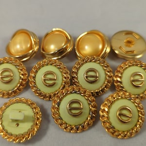 May include: A collection of vintage buttons in various designs. Some buttons feature a gold-tone, pearl-like center with a gold frame. Other buttons have a light green center with a gold rope-like border and a gold letter "e".