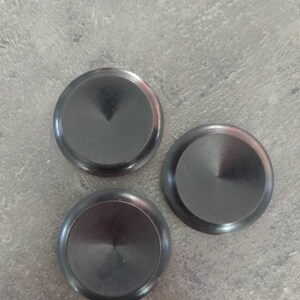 May include: Three black, circular objects with a shiny, metallic appearance are arranged on a gray surface. Each disc has a slightly raised edge and a central, conical indentation, creating a subtle, reflective surface.