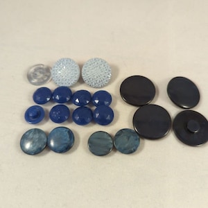 May include: A collection of various buttons in shades of blue, black, and clear. The buttons vary in size, shape, and texture, including faceted, patterned, and smooth designs. Some buttons have a shank, while others have holes for sewing.