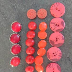 May include: A collection of various red and orange buttons. The buttons include round, oval, and flower-shaped designs. Some buttons have a marbled effect, while others have a shiny, smooth finish. The buttons are arranged on a gray surface.