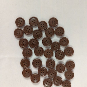 May include: A collection of brown, four-hole buttons scattered on a white surface. The buttons are round with a slightly glossy finish, suitable for sewing projects or garment embellishment.