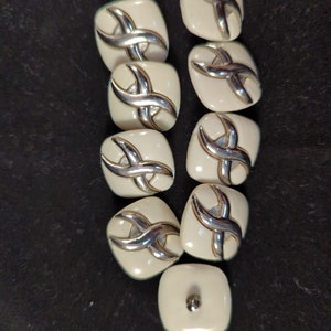 May include: A collection of ten square, off-white buttons with a silver-colored X-shaped design. The buttons have a slightly rounded shape and a small silver button on the back. The buttons are arranged on a dark surface.