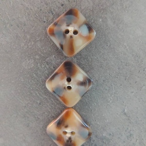 May include: Three diamond-shaped buttons with a marbled pattern in shades of brown, tan, and blue-gray. Each button has two holes for sewing. The buttons are arranged vertically against a gray background.