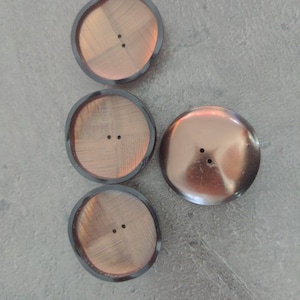 May include: Four copper-colored buttons with two holes each. The buttons have a dark gray rim and a textured surface. One button has a smooth, glossy finish. The buttons are round and vary slightly in shape.