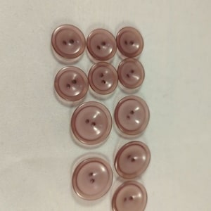 May include: A collection of eleven mauve-colored buttons with two holes each. The buttons are arranged in a vertical pattern on a white surface. The buttons vary slightly in size, with a glossy finish.