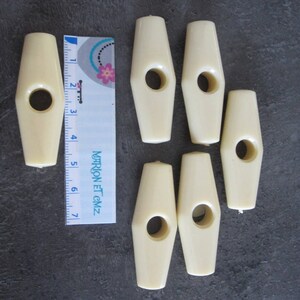 May include: Ivory-colored toggle clasps with a central hole, arranged on a dark surface. A ruler is placed next to the clasps, showing measurements in inches. The clasps have a smooth, rounded shape, suitable for various craft projects.