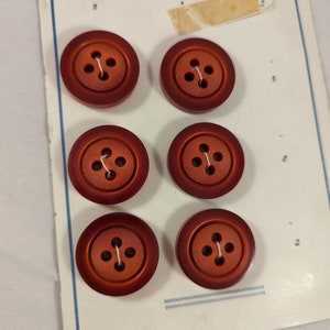 May include: Six round, reddish-brown buttons with four holes each, attached to a vintage card. The buttons have a slightly raised edge and are made of a solid material. The card has a blue border and a small tag.