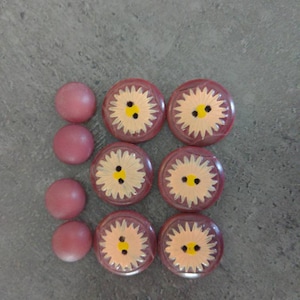 May include: A collection of decorative buttons and beads. The buttons are round with a dark red border and a yellow and white floral design. Three pink beads are also present.