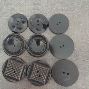 May include: Nine black buttons with various designs. The buttons are round and made of a dark material. Some buttons have floral patterns, while others have geometric or plain designs. Each button has two or four holes for sewing.