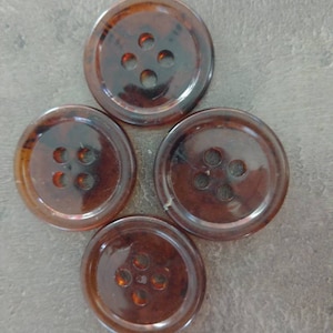 May include: Four round, brown buttons with four holes each, arranged in a cloverleaf pattern. The buttons have a tortoiseshell appearance, with variations in color and translucence. They are likely made of plastic or resin.