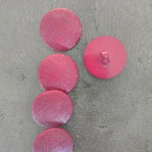 May include: Five round, pink buttons. Four buttons have a textured, woven fabric appearance. One button is solid pink with a shank on the back. The buttons are arranged on a gray surface.