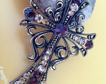May include: A silver-toned cross pendant with intricate detailing and a floral design. The cross is adorned with clear and burgundy-coloured rhinestones.