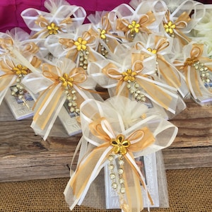 May include: A group of small white and gold gift bags with white and gold ribbon bows. Each bag contains a small silver picture frame with a white and gold design. The bags are sitting on a wooden surface.