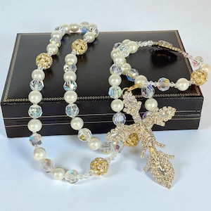 May include: A pearl necklace and bracelet set with gold accents and a sparkling cross pendant. The necklace and bracelet feature white pearls, clear crystal beads, and gold-coloured, rhinestone-studded spheres. The cross pendant is gold-toned and encrusted with clear rhinestones.