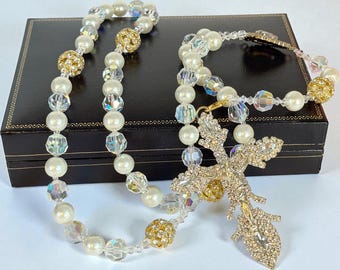 Puede incluir: Un conjunto de collar y pulsera de perlas con detalles dorados y un colgante de cruz brillante. El collar y la pulsera presentan perlas blancas, cuentas de cristal transparente y esferas doradas con incrustaciones de pedrería. El colgante de cruz es dorado y está incrustado con pedrería transparente.