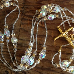 May include: A white crystal rosary with gold accents and a gold crucifix. The rosary beads are faceted and iridescent.