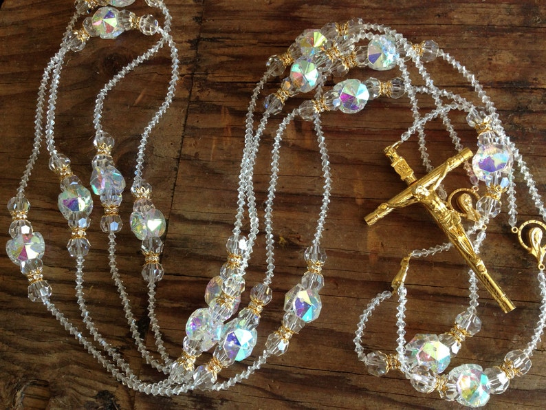 May include: A white crystal rosary with gold accents and a gold crucifix. The rosary beads are faceted and iridescent.