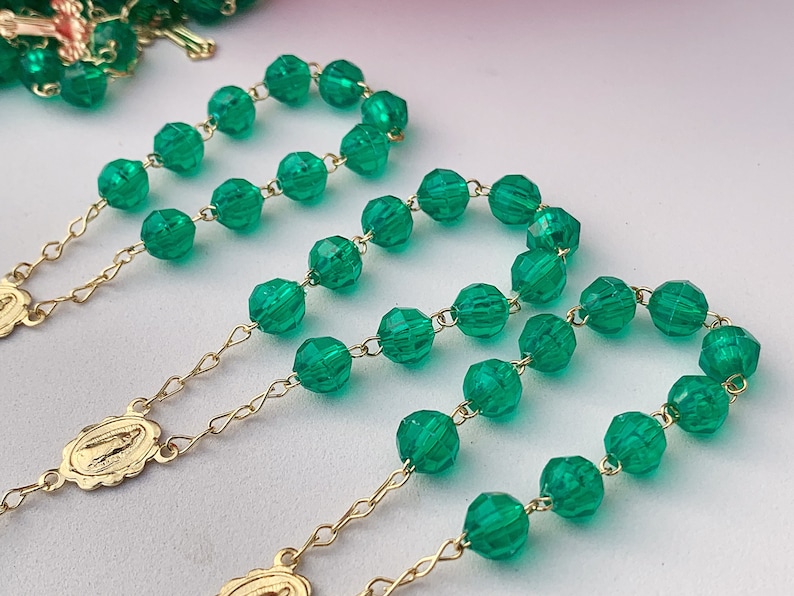 Puede incluir: Tres rosarios de cuentas de vidrio verde con cadenas y medallones de oro. Los rosarios est&aacute;n dispuestos sobre una superficie blanca.