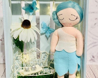 Puede incluir: Una muñeca de fieltro hecha a mano, con cabello azul y falda celeste, se encuentra en una caja de madera celeste. La caja contiene un banco blanco en miniatura, una gran flor blanca y mariposas azules. Luces de hadas añaden un toque mágico.