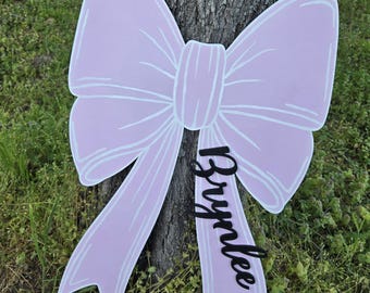 Puede incluir: Un gran lazo rosa con ribete blanco y el nombre "Brynnlee" escrito en letras negras. El lazo está hecho de madera y es un letrero decorativo.