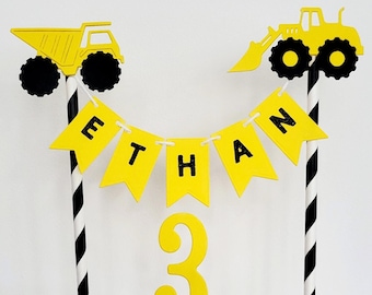 May include: Yellow and black construction vehicle cake topper with a banner that says "ETHAN" and a number "3" on a stick.