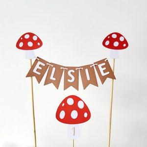 May include: A cake topper with a brown banner that says "ELSIE" and red and white mushroom decorations. The banner is strung between two mushroom toppers. There is a red and white mushroom with a white number "1" on the bottom.