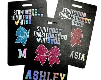 May include: Three black ID badge holders with a holographic finish. Each holder features a glittery bow, the words "STUNT HARD TUMBLE STRONG HIT ZERO" and a name. The names are "M", "ASHLEY", and "ASIA".
