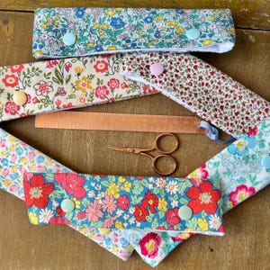 May include: Several floral fabric headbands in shades of blue, red, and yellow, secured with buttons. A copper ruler and small scissors are also visible. The items are arranged on a wooden surface.