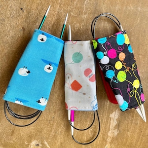 May include: Three fabric circular knitting needle cases. The left case is bright blue with white sheep. The centre case is light grey with colourful yarn balls and needles. The right case is black with a pattern of vibrant, multi-coloured yarn balls.