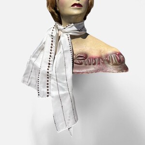 May include: White silk scarf with brown polka dots and thin stripes. The scarf is tied around a mannequin's neck.