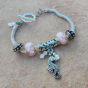 Pode incluir: Um bracelete de prata com uma corrente de prata e um fecho de cavalo-marinho de prata. O bracelete também tem contas de vidro rosa e branco com desenhos florais.