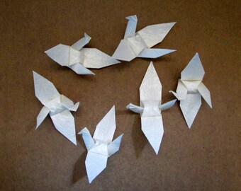 May include: Five white origami cranes folded from paper. The cranes are arranged in a starburst pattern on a brown surface.