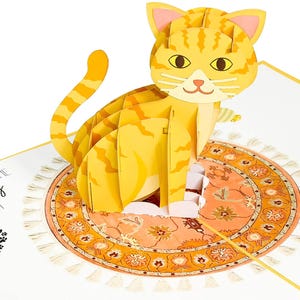 May include: A 3D pop-up card featuring a yellow tabby cat sitting on a colorful rug with fringe. The card is open and the cat is popping up from the card.