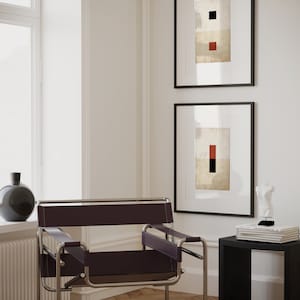 Op de afbeelding: Een zwarte leren en chromen stoel met een zwarte bijzettafel en twee ingelijste abstracte kunstprints met geometrische vormen in rood en zwart op een witte achtergrond.