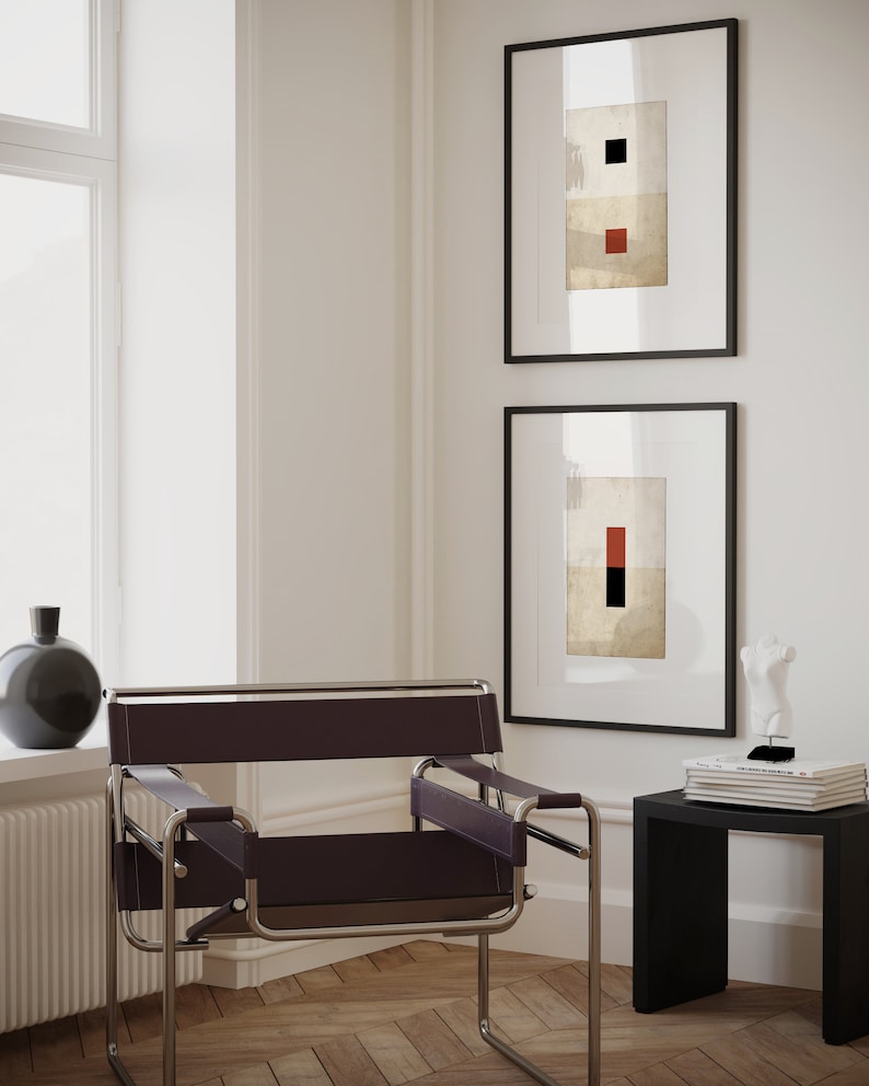 Op de afbeelding: Een zwarte leren en chromen stoel met een zwarte bijzettafel en twee ingelijste abstracte kunstprints met geometrische vormen in rood en zwart op een witte achtergrond.