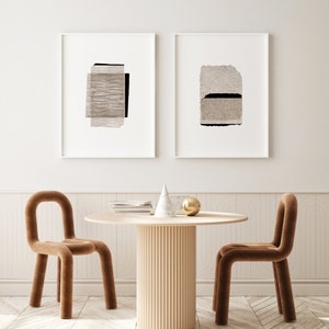 May include: Two framed abstract art prints with beige and black paper textures hang on a white wall above a round wooden table with a fluted base. Two brown velvet chairs with rounded backs are placed on either side of the table.