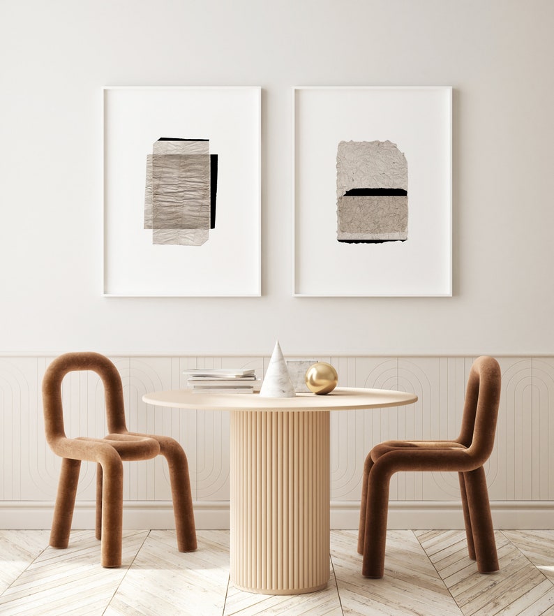 May include: Two framed abstract art prints with beige and black paper textures hang on a white wall above a round wooden table with a fluted base. Two brown velvet chairs with rounded backs are placed on either side of the table.