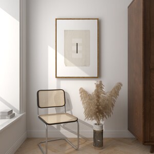 Op de afbeelding: Een minimalistische zwart-wit abstracte kunstprint in een houten lijst hangt aan een witte muur. Een stoel van geweven riet staat voor de kunstprint. Een vaas met gedroogd pampasgras staat op de vloer naast de stoel.