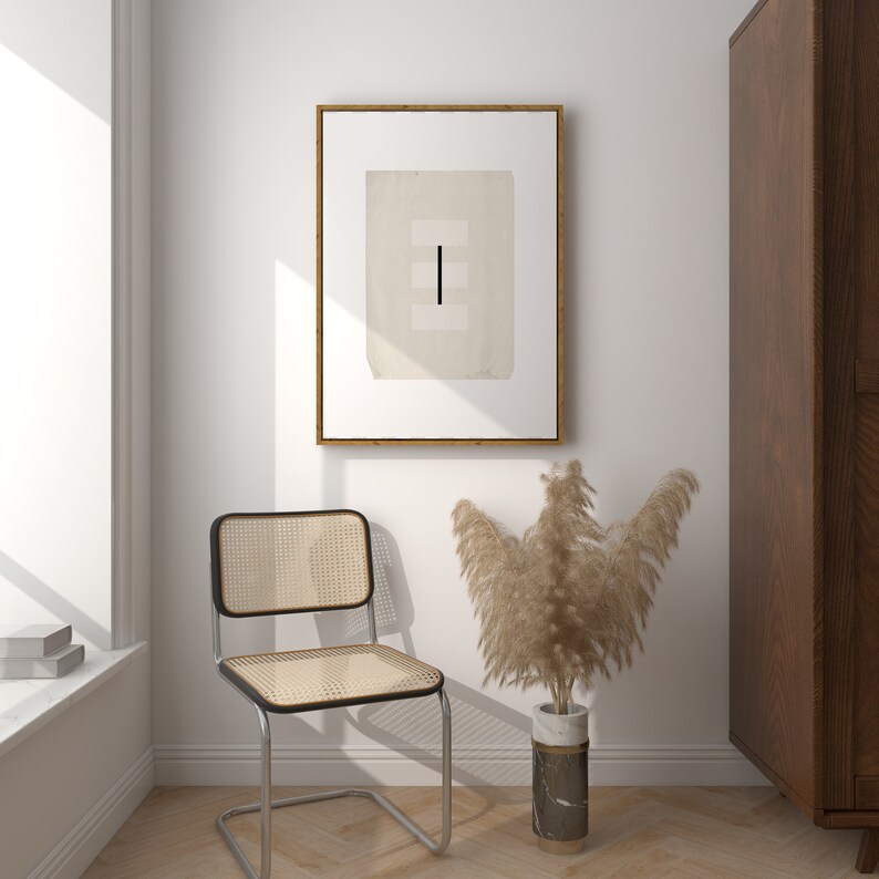 Op de afbeelding: Een minimalistische zwart-wit abstracte kunstprint in een houten lijst hangt aan een witte muur. Een stoel van geweven riet staat voor de kunstprint. Een vaas met gedroogd pampasgras staat op de vloer naast de stoel.