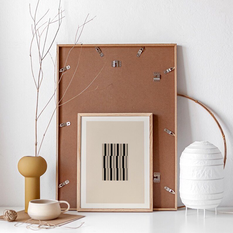 May include: A brown wooden frame with a white mount and a black and white geometric print inside. The frame is hanging on a white wall with a vase of branches in the foreground.