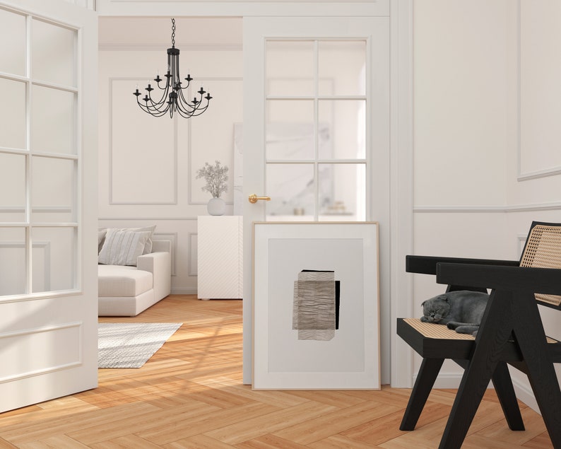 May include: A white room with a black chair and a black and white abstract print hanging on the wall. A cat is sleeping on the chair. The room has white walls and a wooden floor.